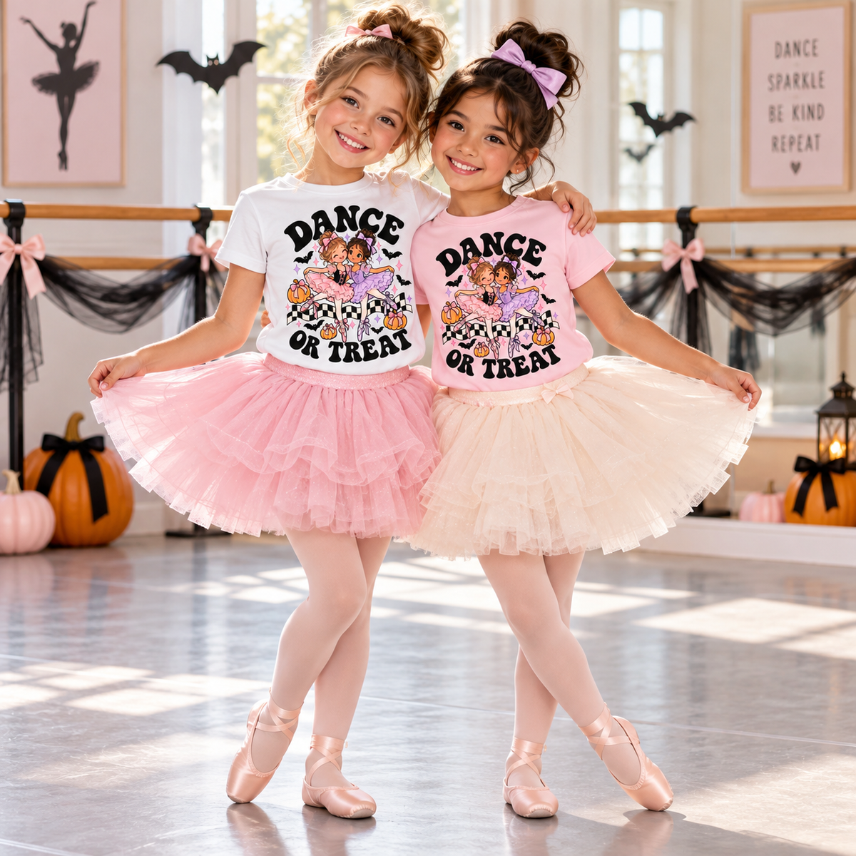 Two young ballerina best friends modeling Dance or Treat Halloween ballet T-shirts with pink and lavender tutu dancer graphics, pumpkins, bats, and bows. Girls wear pink and white shirts with fluffy tutus and ballet slippers in a Halloween-decorated dance studio. Cute girls’ Halloween dance class outfit.