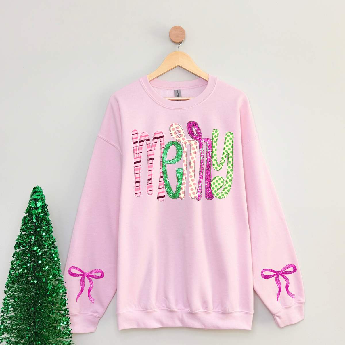 Light pink Merry Christmas crewneck sweatshirt with colorful pink and green patterned lettering, faux glitter details, and pink bow graphics on both sleeves\. Preppy coquette Christmas sweatshirt for women\.