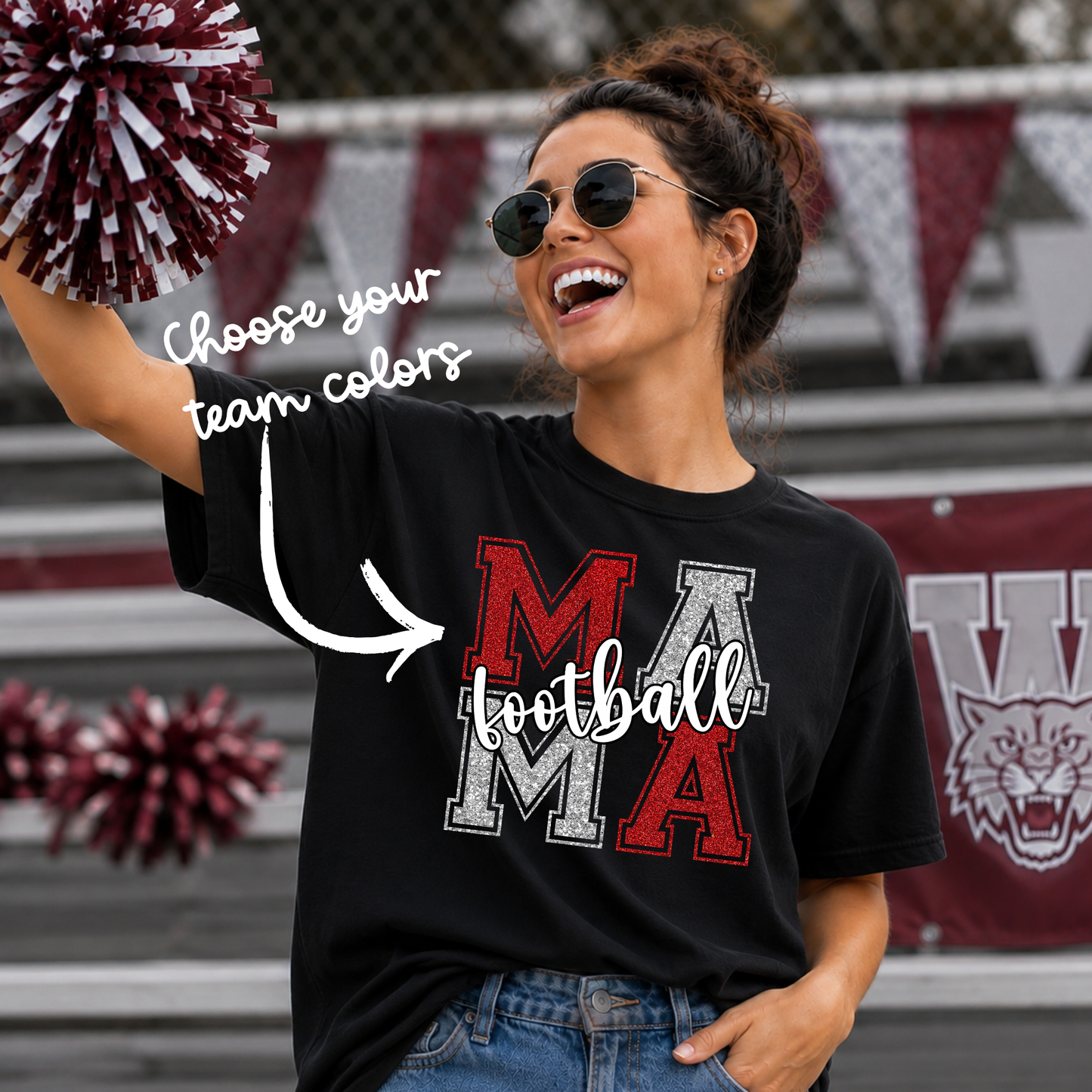 A woman wearing a black graphic t-shirt with 'MAMA football' printed on the front in red, white, and gray. She is holding a maroon and white pom-pom and standing in front of stadium bleachers with banners in the background.