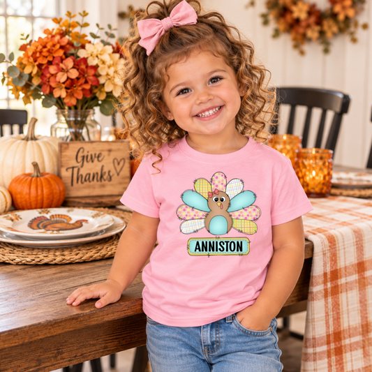Toddler girl with curly hair wearing a personalized light pink Thanksgiving T-shirt featuring a colorful pastel patchwork turkey and custom name Anniston, standing beside a decorated Thanksgiving dining table.