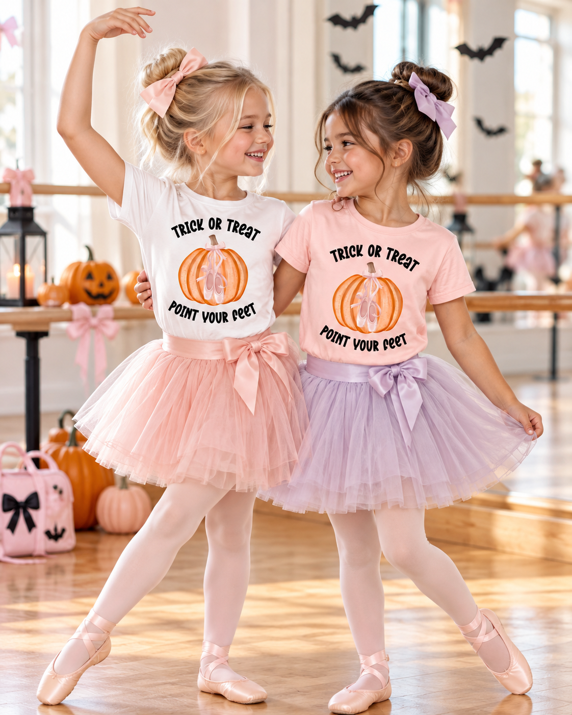 Two young ballerina best friends modeling Trick or Treat Point Your Feet Halloween ballet T-shirts in white and pink, paired with pink and lavender tutus and ballet slippers. Shirts feature an orange pumpkin with pink ballet shoes and bow, perfect for girls’ Halloween dance class outfits.