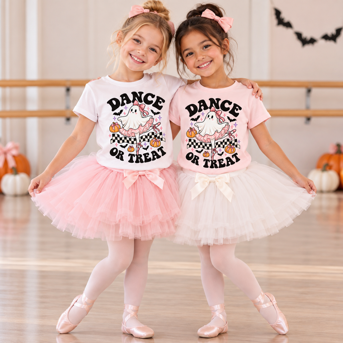 Two young ballerina best friends wearing Dance or Treat Halloween ballet T-shirts in white and light pink, paired with fluffy pink and cream tutus and ballet slippers. Shirts feature a cute ghost ballerina with pink bows, pumpkins, bats, and checkerboard accents. Girls pose together in a dance studio.
