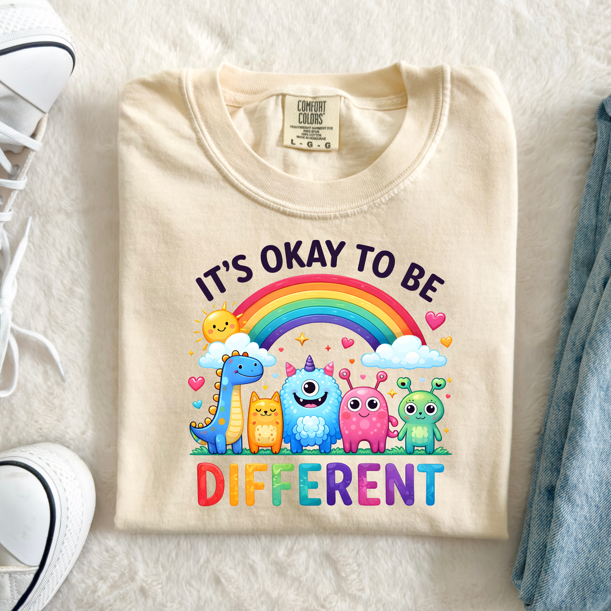 Comfort Colors ivory teacher T-shirt featuring a colorful rainbow, cute smiling monsters, and It's Okay to Be Different lettering, perfect for special education teachers, school counselors, and inclusive classrooms.