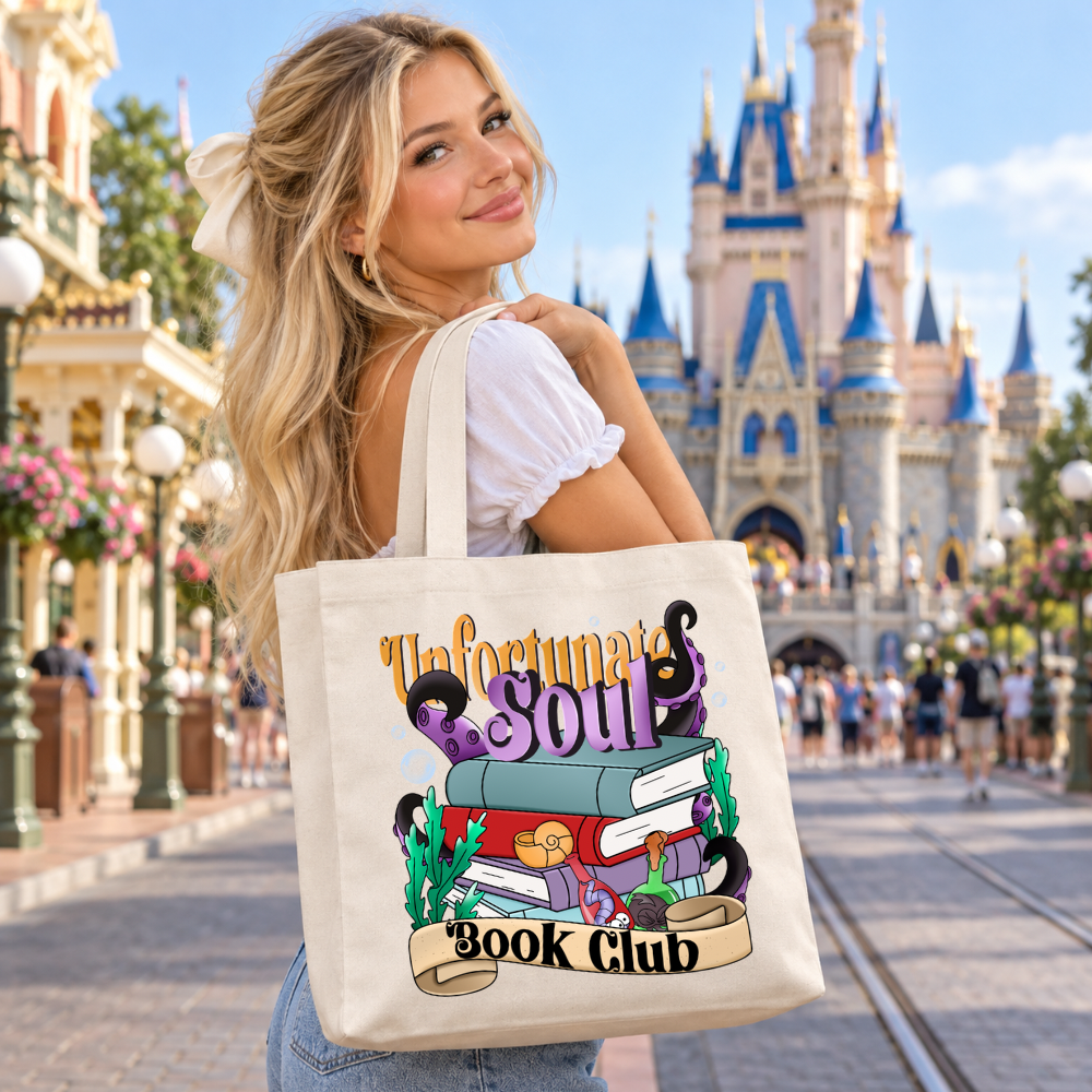 Blonde woman carrying a natural canvas tote bag featuring a colorful Unfortunate Souls Book Club design with books and purple sea-inspired artwork in front of a magical theme park castle.